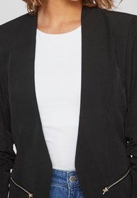 Černý blazer s moderním střihem, dlouhými rukávy, otevřeným předním dílem a zlatými zipy na bocích. Nositý přes bílou košili.