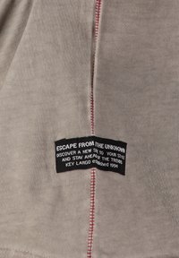 Tejido beige con una superficie texturizada, con una etiqueta de fondo negro y texto blanco, cosida con hilo rojo a lo largo de la costura.