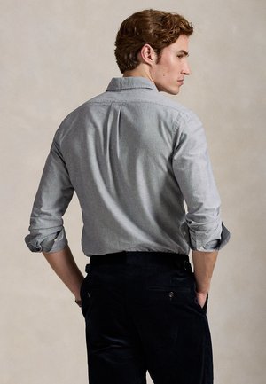 Férfi világosszürke hosszú ujjú, gombos ingben, felhajtott ujjakkal és sötét nadrágban, zsebre tett kézzel áll, háttal néz.