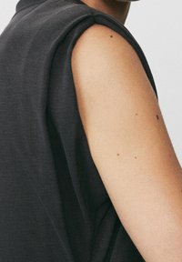 Personne portant un haut noir sans manches, montrant le haut du bras avec plusieurs petits grains de beauté sur un fond clair.