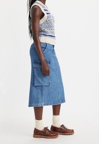 Pletený bezrukávový svetr s modro-krémovými vzory, doplněný modrou džínsovou sukní s velkou boční kapsou a hnědými šněrovacími botami.