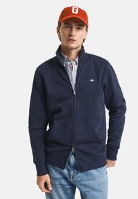 Felpa zip-up di colore blu navy in cotone, caratterizzata da un logo discreto sul petto, con due tasche frontali e un colletto alto.