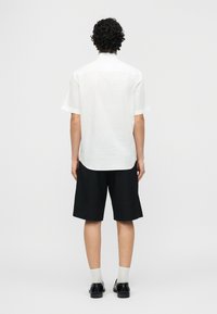 Άνδρας με σγουρά μαλλιά στέκεται με την πλάτη γυρισμένη φορώντας άσπρο πουκάμισο με κοντά μανίκια, μαύρο σορτς, λευκές κάλτσες και μαύρα παπούτσια επίσημου τύπου σε απλό φόντο.