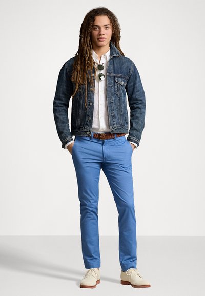 Giubbotto di jeans con vestibilità strutturata, camicia bianca con bottoni, chinos blu e scarpe chiare; gli accessori includono una cintura e occhiali da sole.