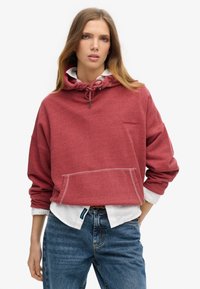 Sudadera con capucha roja y acabado texturizado, con cordón ajustable. Presenta un bolsillo frontal y un sutil detalle de logo. Llevada sobre una camiseta blanca.