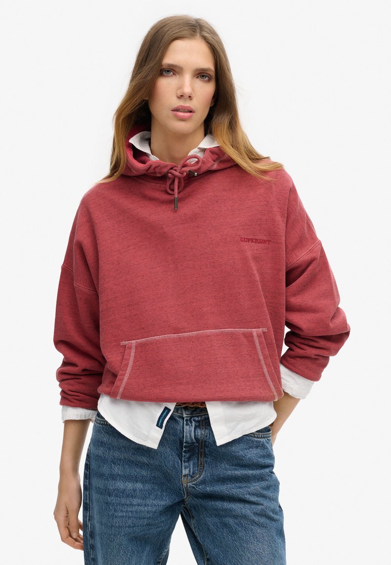 Sudadera con capucha roja y acabado texturizado, con cordón ajustable. Presenta un bolsillo frontal y un sutil detalle de logo. Llevada sobre una camiseta blanca.