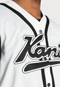 Bílý baseballový dres s černými akcenty a knoflíky, zdobený výrazným logem "Kani" v tučné černé výšivce přes hrudník.