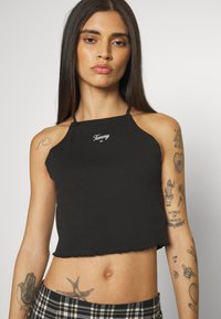 Black cropped tank top with a rounded neckline and frayed hem. Features a white "Tommy" logo on the chest. Smooth texture, fitted design.