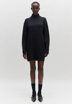 Zwarte oversized lange mouw jurk met een hoge halslijn. Gemaakt van zachte, gestructureerde stof en een rechte snit, gecombineerd met zwarte enkelsokken en puntige schoenen.