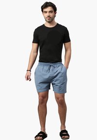 T-shirt in cotone nero, pantaloni corti blu chiaro con vita elastica e cordoncino, abbinati a sandali neri con suola piatta, indossati da una figura maschile.