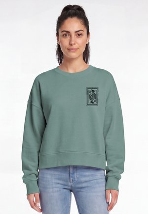 Femme aux cheveux foncés attachés en queue de cheval, portant un sweat-shirt vert clair avec un motif de carte à jouer noire sur le haut gauche devant, et un jean bleu clair.