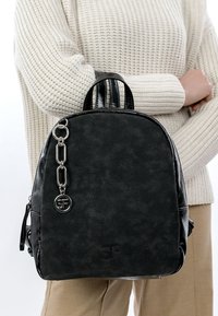 Svart ryggsäck av texturerat syntetmaterial med dubbla handtag, en silverkedja som detalj och en cirkulär logotyp.