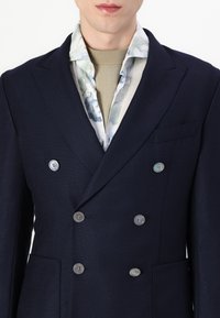 Blazer doppiopetto navy in tessuto testurizzato con bottoni argentati, con colletto a fiori che spunta da sotto.