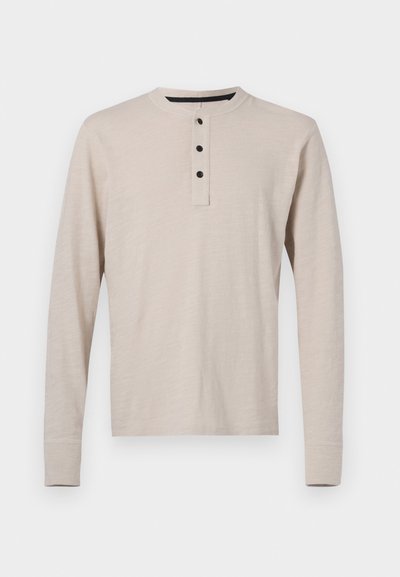 T-shirt henley à manches longues en beige clair, fabriqué en tissu doux. Il possède un col rond et trois boutons noirs le long de l'ouverture.