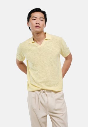 Jeune homme aux cheveux noirs portant une chemise jaune pâle à manches courtes et un pantalon beige clair avec un cordon, debout devant un fond clair uni.