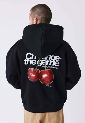 Uomo che indossa felpa nera con cappuccio con grandi ciliegie rosse e scritta "Change the game" sulla schiena, pantaloni beige, di spalle.