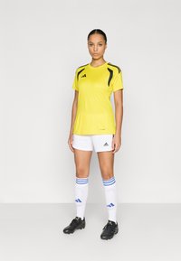 Κίτρινο αθλητικό μπλουζάκι με μαύρες λεπτομέρειες, λευκό σορτς και ριγωτές κάλτσες. Μαύρα ποδοσφαιρικά παπούτσια ολοκληρώνουν το σύνολο αθλητικής ένδυσης.