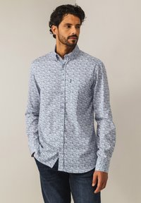 Langärmliges, button-down Hemd in Hellblau mit floralem Muster, ausgestattet mit einem klassischen Kragen und einer Brusttasche. Getragen mit dunklen Jeans.