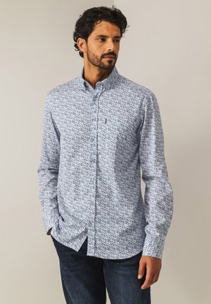 Langärmliges, button-down Hemd in Hellblau mit floralem Muster, ausgestattet mit einem klassischen Kragen und einer Brusttasche. Getragen mit dunklen Jeans.