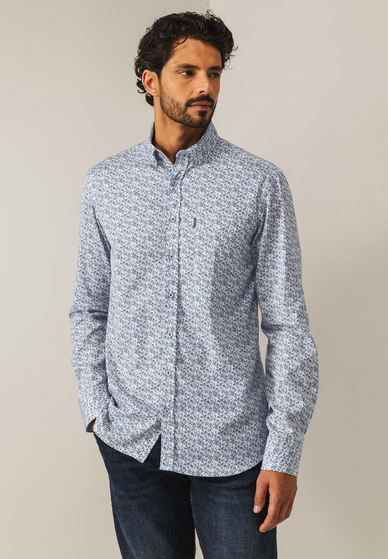 Langärmliges, button-down Hemd in Hellblau mit floralem Muster, ausgestattet mit einem klassischen Kragen und einer Brusttasche. Getragen mit dunklen Jeans.