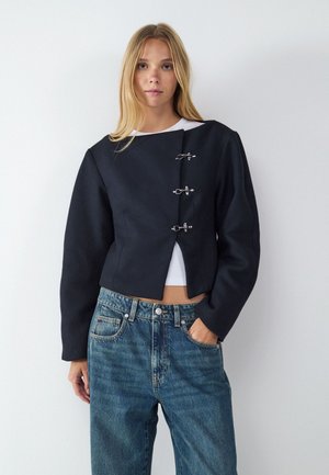 Lyshåret kvinde iført en sort cropped jakke med tre sølvspænder over en hvid top og højtaljede blå jeans, stående mod en hvid væg.