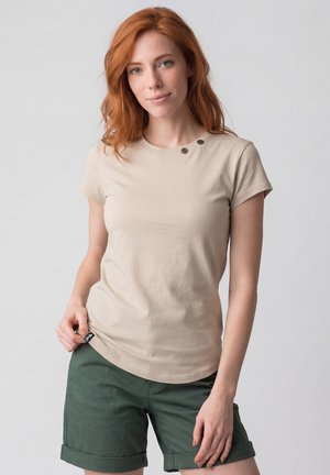 Junge Frau mit roten Haaren, die ein beiges kurzärmeliges Hemd mit Knöpfen und grüne hochgekrempelte Shorts trägt und vor einem schlichten Hintergrund steht.