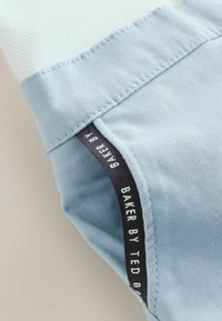 Sezione del capo azzurro chiaro con parte superiore bianca e strutturata, con un'etichetta nera con testo bianco. Tessuto liscio con dettagli di cuciture visibili.