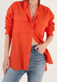 Femme portant une chemise orange vif à boutons avec les manches retroussées et un jean bleu clair, la main dans la poche, plusieurs colliers en or superposés visibles.