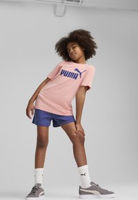 Camiseta de algodón rosa con el logotipo "PUMA" en color marino, combinada con pantalones cortos en color marino. El modelo lleva zapatillas grises y calcetines blancos con logotipo negro.