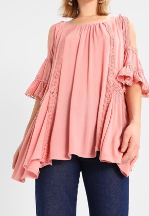 Rosa Bluse mit kalten Schultern, Rüschensäumen, Spitzenverzierungen und lockerer Passform. Getragen mit blauen Denim-Jeans. Weicher, leichter Stoff.