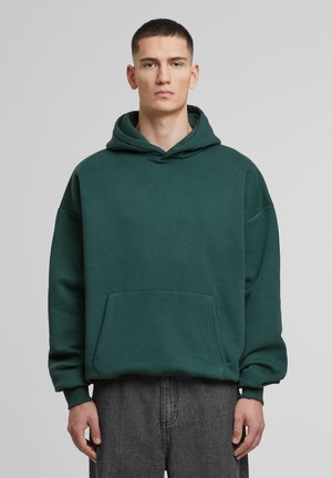 Dark green oversized hoodie made of soft material, featuring a front pocket and a drawstring hood. Model wears gray pants.