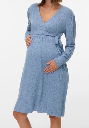 Femme enceinte en robe portefeuille bleu clair à manches longues, tenant délicatement son ventre devant un fond blanc uni.