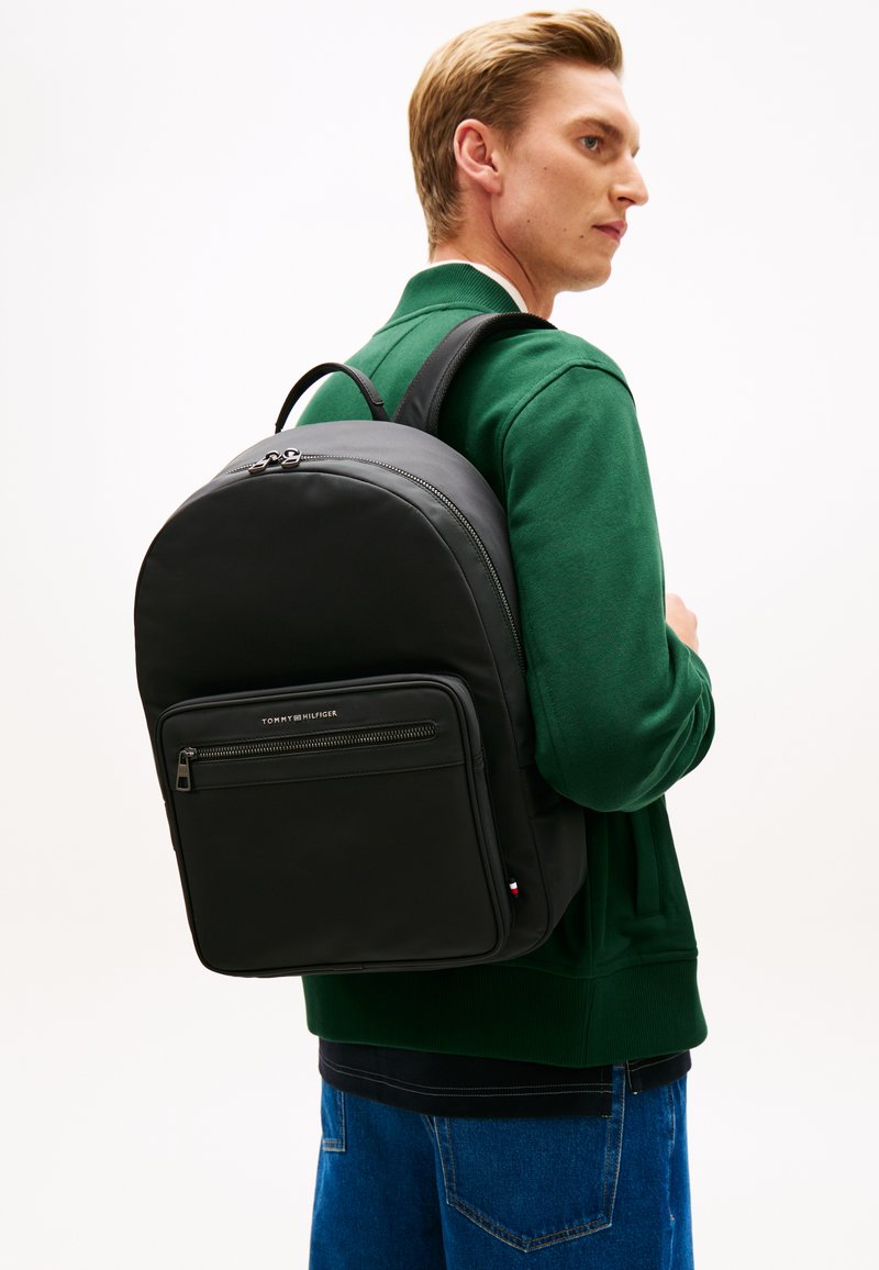 Νέος άνδρας που φοράει πράσινο φούτερ και μπλε τζιν κουβαλάει μια μαύρη τσάντα πλάτης Tommy Hilfiger στην πλάτη του, στραμμένος προς τα δεξιά.