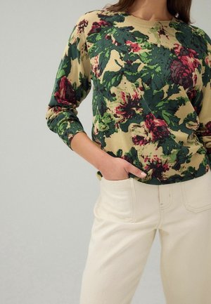 Žena v béžových kalhotách a dlouhém svetru s květinovým vzorem v zelené, červené a béžové barvě, s jednou rukou v kapse.