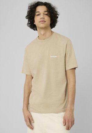 Jeune homme aux cheveux bouclés portant un t-shirt beige à manches courtes avec le texte « Cœur de pierre » sur la poitrine, sur un fond gris uni.