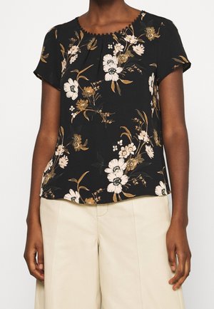 Femme portant un chemisier noir à manches courtes avec des motifs floraux beige et blancs et un pantalon large crème.