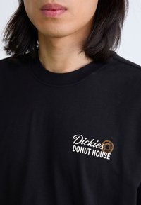 Crna pamučna majica s bijelim i smešnim logotipom koji glasi "Dickies DONUT HOUSE," vidljivim na gornjoj lijevoj strani, s okruglim izrezom.
