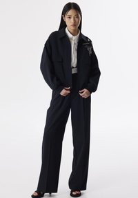 Veste oversize marine avec poches à rabat, ornée de chaînes, associée à un pantalon ajusté taille haute et des sandales à plateforme noires.