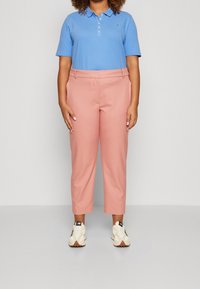 Lichtblauwe polo met knopen, gecombineerd met roze op maat gemaakte broeken. Schoenen zijn wit en zwarte sneakers. Eenvoudige, casual outfit.