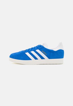 Zapatilla de ante azul con suela de goma blanca, que presenta tres rayas blancas y "Gazelle" en dorado en el lateral. Diseño clásico de corte bajo.