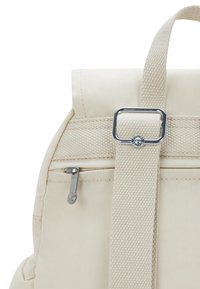 Mochila de lona beige que cuenta con un bolsillo con cremallera, una correa prominente con una hebilla plateada y detalles de tela texturizada.