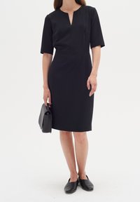 Svart kortärmad klänning med V-ringning, figurnära silhuett och knälång fåll. Bärs med en svart handväska och platta skor.