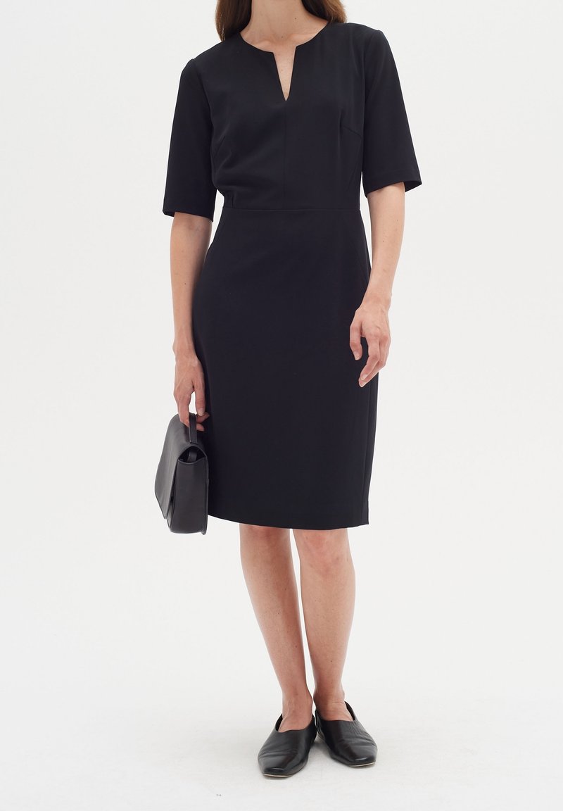 Svart kortärmad klänning med V-ringning, figurnära silhuett och knälång fåll. Bärs med en svart handväska och platta skor.