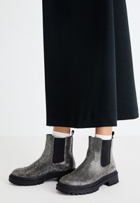 Bottines avec des côtés élastiques noirs et des dessus argentés pailletés, portées avec des chaussettes blanches et une longue jupe noire.