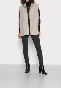 Chaleco sin mangas beige con dos bolsillos frontales, cierre de botón, usado sobre un suéter negro estampado y combinado con jeans oscuros y botas de tacón cuadrado.