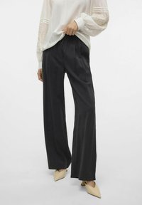 Zwarte, hoge taille, wijde broek met plooien. De stof heeft een gladde textuur. Gepaard met een witte blouse en lichte, puntige schoenen.