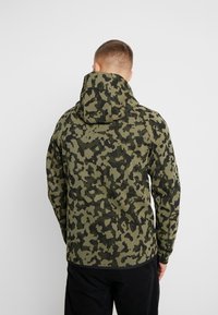 Maskovací mikina v odstínech zelené a černé, s vysokým límcem a kapucí. Vyrobena z texturované látky, s volným střihem a dlouhými rukávy.