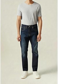 Tmavě modré džínové kalhoty se střihm přímo do nohy, pěti kapsami a kontrastním šitím, doplněné světle šedým tričkem s kulatým výstřihem a bílými nazouvacími botami.