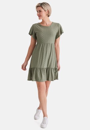 Frau läuft und trägt ein olivgrünes Spitzenkleid mit kurzen Ärmeln und Rüschensaum sowie weiße Turnschuhe vor einem schlichten Hintergrund.