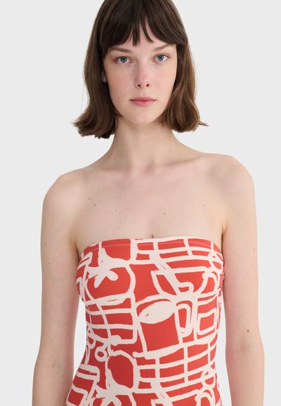 Jeune femme aux cheveux bruns courts portant une robe rouge sans bretelles ornée de formes et de lignes blanches abstraites sur un fond clair uni.